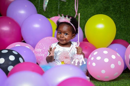 A little girl wearing a tiara celebrates her birthday among colorful balloons